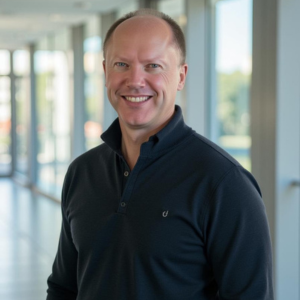 Smiling man in a dark long-sleeve shirt standing in a bright hallway with large windows.